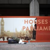 A rough sleeper lies on a suitcase in front of an advertisement board showing the Houses of Parliament in central London